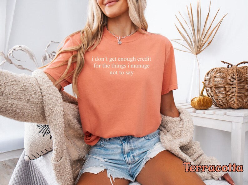 K&ouml;nnte beinhalten: Terracottafarbenes T-Shirt mit dem wei&szlig;en Text "I don't get enough credit for the things I manage not to say". Das Shirt wird mit Jeansshorts und einer cremefarbenen Strickjacke kombiniert. Dekorative K&uuml;rbisse und ein Weidenkorb sind im Hintergrund.