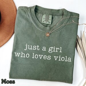 以下が含まれることがあります： 前面に白い文字で「just a girl who loves viola」とプリントされたモスグリーンの半袖Tシャツ。クルーネックで、襟の内側にはComfort Colorsのブランドラベルがあり、サイズSであることが示されています。