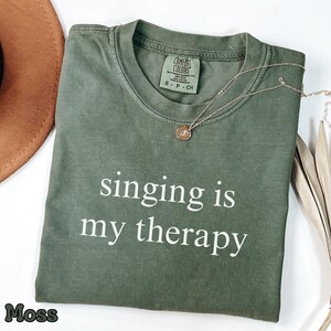 Op de afbeelding: Een mosgroen T-shirt met korte mouwen, een ronde hals en de witte tekst "singing is my therapy" op de voorzijde. Het shirt heeft een Comfort Colors merklabel in de nek, wat maat S aangeeft, en is gemaakt van katoen.