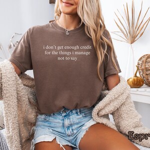 K&ouml;nnte beinhalten: Espressofarbenes T-Shirt mit dem wei&szlig;en Text "I don't get enough credit for the things I manage not to say". Das Shirt wird mit Jeansshorts kombiniert. Ein cremefarbener Cardigan ist &uuml;ber den Schultern drapiert.