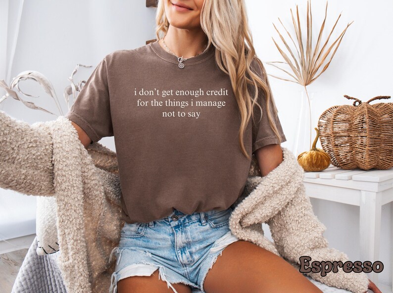 K&ouml;nnte beinhalten: Espressofarbenes T-Shirt mit dem wei&szlig;en Text "I don't get enough credit for the things I manage not to say". Das Shirt wird mit Jeansshorts kombiniert. Ein cremefarbener Cardigan ist &uuml;ber den Schultern drapiert.