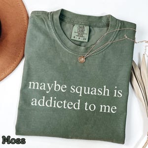 Könnte beinhalten: Ein moosgrünes Kurzarm-T-Shirt mit Rundhalsausschnitt und dem weißen Text "maybe squash is addicted to me" auf der Vorderseite. Das T-Shirt hat ein Comfort Colors Markenetikett am inneren Halsausschnitt mit der Größenangabe S-P-CH.