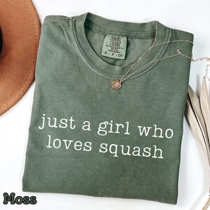 May include: A moss green short-sleeved t-shirt with a crew neckline and the white text "just a girl who loves squash" printed on the front. The shirt features a Comfort Colors brand label at the inner neck and is made of 100% cotton fabric.