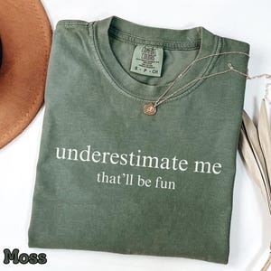 May include: A moss green t-shirt with the phrase "underestimate me that'll be fun" printed in white. A gold necklace with a pendant rests on the shirt. The shirt is folded and placed on a white surface.