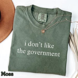 May include: Moss green t-shirt with the text "i don't like the government" in white. A gold necklace with a pendant is on the shirt. A brown hat and dried plant are in the background.