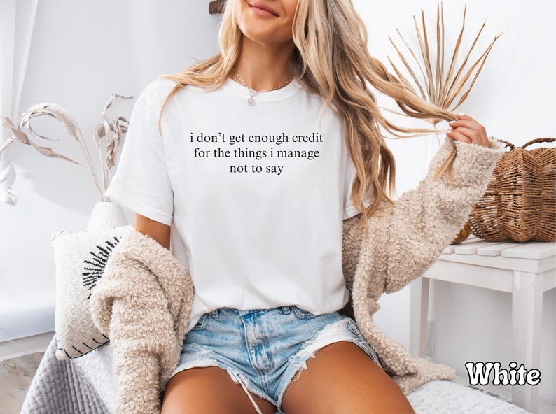 K&ouml;nnte beinhalten: Wei&szlig;es T-Shirt mit dem Text "i don't get enough credit for the things i manage not to say" in Schwarz. Das Model tr&auml;gt Jeansshorts und eine beige Jacke. Ein wei&szlig;es Dekokissen und ein Weidenkorb sind im Hintergrund.