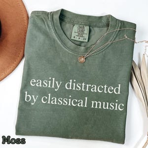 Pode incluir: Camiseta verde musgo com o texto branco "easily distracted by classical music". Um colar de ouro com um pequeno pingente repousa sobre a camiseta dobrada sobre uma superfície branca.