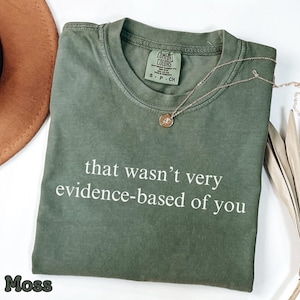 Puede incluir: Una camiseta verde musgo con la frase "that wasn't very evidence-based of you" en blanco. Un collar dorado con un colgante está drapeado sobre la camiseta. La camiseta está doblada y sobre una superficie blanca.