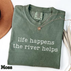 May include: A moss green t-shirt with the text "life happens the river helps" in white. A gold necklace with a pendant is on the shirt. A brown hat and dried leaves are in the background.