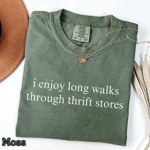 May include: Moss green t-shirt with the text "i enjoy long walks through thrift stores" in white. A gold necklace with a pendant is on the shirt. The shirt is folded and has a Comfort Colors tag.