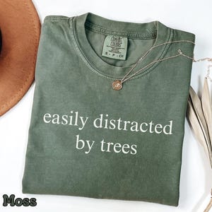 Op de afbeelding: Mosgroen t-shirt met de witte tekst "easily distracted by trees". Een goudkleurige ketting met een kleine hanger ligt op het shirt. Een bruine hoed en een gedroogde plant staan op de achtergrond.