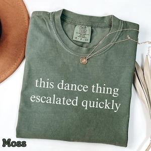 Puede incluir: Camiseta verde musgo con la frase blanca "this dance thing escalated quickly". Un collar dorado con un colgante descansa sobre la camiseta. Un sombrero marrón y hojas de palma secas están en el fondo.