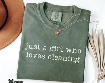 May include: Moss green t-shirt with the text "just a girl who loves cleaning" in white. A gold necklace with a small pendant rests on the shirt. A brown hat and dried palm leaves are in the background.
