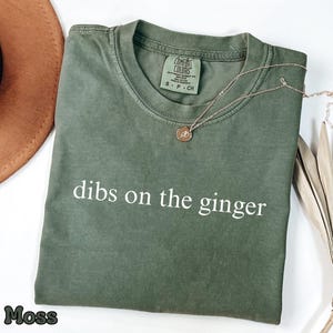 May include: A moss green t-shirt with the text "dibs on the ginger" in white. A gold necklace with a pendant is on the shirt. The shirt is folded on a white surface. The t-shirt is a Comfort Colors brand.