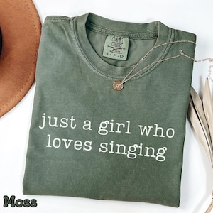 Op de afbeelding: Een mosgroen T-shirt met korte mouwen en een ronde hals. Het shirt heeft de witte opdruk "just a girl who loves singing" op de borst. Een klein label in de kraag toont de tekst "Comfort Colors" en de maat "S-P-CH" is zichtbaar.