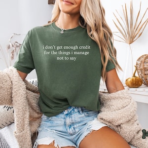 K&ouml;nnte beinhalten: Moosgr&uuml;nes T-Shirt mit dem wei&szlig;en Text "i don't get enough credit for the things i manage not to say". Das Shirt wird mit Jeansshorts und einer beigen Strickjacke getragen. Das Wort "Moss" steht unten rechts.