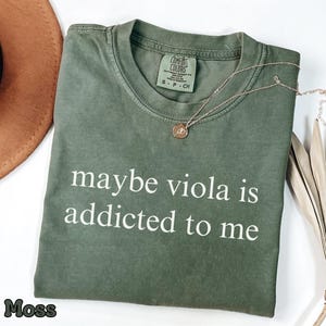 May include: A moss green short-sleeved t-shirt with a crew neckline and the white text "maybe viola is addicted to me" printed on the front. The shirt features a Comfort Colors brand label at the inner neck and is made of 100% cotton fabric.