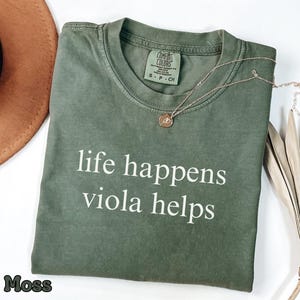 May include: A moss green short-sleeve t-shirt with a crew neckline. The shirt features the white text "life happens viola helps" printed on the front. A small tag inside the collar reads "Comfort Colors" with size "S - P - CH" visible.