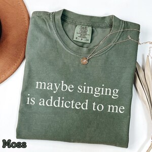 Puede incluir: Una camiseta de manga corta color verde musgo con cuello redondo y etiqueta de la marca Comfort Colors. La prenda presenta el texto impreso en blanco 'maybe singing is addicted to me' centrado en el pecho. Un collar dorado descansa sobre la tela.