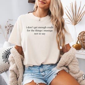 K&ouml;nnte beinhalten: Cremefarbenes T-Shirt mit dem Text "i don't get enough credit for the things i manage not to say" in Schwarz. Das Shirt wird mit Jeansshorts getragen. Das Wort "Ivory" steht in einer Schreibschrift unten rechts.