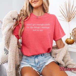 K&ouml;nnte beinhalten: Ein wasserfarbenes T-Shirt mit dem Text "i don't get enough credit for the things i manage not to say" in Wei&szlig;. Das Shirt wird mit Jeansshorts kombiniert. Das Bild enth&auml;lt auch dekorative K&uuml;rbisse und einen beigen Pullover.