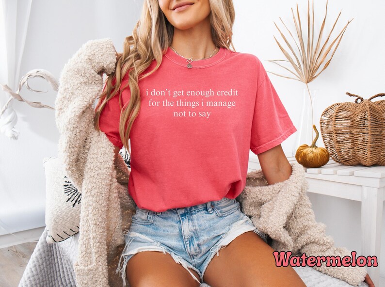 K&ouml;nnte beinhalten: Ein wasserfarbenes T-Shirt mit dem Text "i don't get enough credit for the things i manage not to say" in Wei&szlig;. Das Shirt wird mit Jeansshorts kombiniert. Das Bild enth&auml;lt auch dekorative K&uuml;rbisse und einen beigen Pullover.