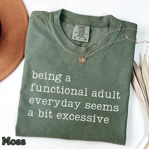 Könnte beinhalten: Moosgrünes T-Shirt mit dem weißen Text "being a functional adult everyday seems a bit excessive". Eine goldene Halskette mit Anhänger liegt auf dem T-Shirt. Ein brauner Hut ist im Hintergrund zu sehen.