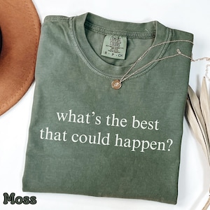 May include: A moss green t-shirt with the text "what's the best that could happen?" in white. A gold necklace with a pendant rests on the shirt. The shirt is folded and placed on a white surface.