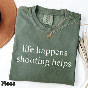 Può includere: Una maglietta verde muschio con la scritta bianca "life happens shooting helps". Una collana d'oro con un piccolo ciondolo è appoggiata sulla maglietta. Un cappello marrone è visibile in alto a sinistra.
