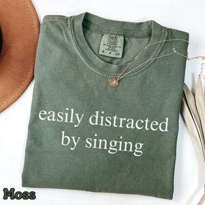 Op de afbeelding: Een mosgroen t-shirt met korte mouwen, een ronde hals en de witte tekst "easily distracted by singing" op de voorkant. Het shirt is voorzien van een Comfort Colors merklabel aan de binnenkant van de kraag met maat S.