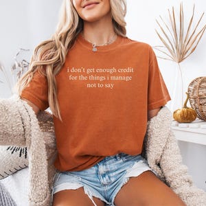 K&ouml;nnte beinhalten: Rostfarbenes T-Shirt mit dem wei&szlig;en Text "I don't get enough credit for the things I manage not to say". Das Shirt wird mit Jeansshorts und einer cremefarbenen Strickjacke kombiniert. Das Bild hat eine warme, herbstliche &Auml;sthetik.