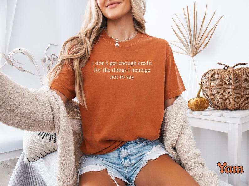 K&ouml;nnte beinhalten: Rostfarbenes T-Shirt mit dem wei&szlig;en Text "I don't get enough credit for the things I manage not to say". Das Shirt wird mit Jeansshorts und einer cremefarbenen Strickjacke kombiniert. Das Bild hat eine warme, herbstliche &Auml;sthetik.