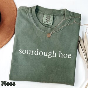 May include: A moss green t-shirt with the words "sourdough hoe" printed in white. A gold necklace with a small pendant rests on the shirt. A brown hat and dried palm leaves are in the background.