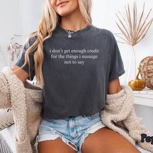 K&ouml;nnte beinhalten: Dunkelgraues T-Shirt mit dem wei&szlig;en Text "i don't get enough credit for the things i manage not to say". Das Shirt wird mit Jeansshorts und einer cremefarbenen Strickjacke kombiniert. Das Wort "Pepper" steht unten rechts.