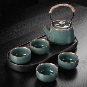 May include: A teal ceramic tea set with a teapot, four small cups, and a serving tray. The teapot has a rope handle and a lid. The cups and teapot have a crackle glaze and a brown rim. The set is on a black tray.