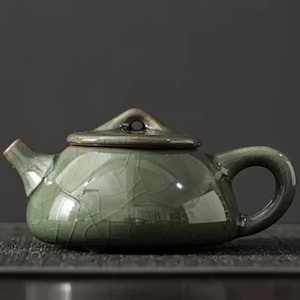 May include: A small, green ceramic teapot with a crackle glaze. The teapot has a rounded body, a curved handle, and a spout. The lid has a small loop. The teapot is designed for brewing tea, with a capacity of approximately 200ml.