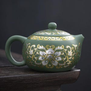 May include: A green ceramic teapot with a floral design in silver and gold. The teapot has a rounded body, a curved handle, and a lid with a small knob. The floral pattern is centered on the body and lid.