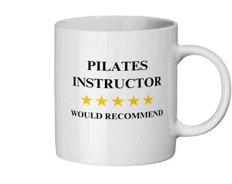 Op de afbeelding: Witte keramische mok met een gebogen handvat. De mok heeft de tekst "PILATES INSTRUCTOR WOULD RECOMMEND" in het zwart, met vijf gouden sterren onder de woorden "INSTRUCTOR".
