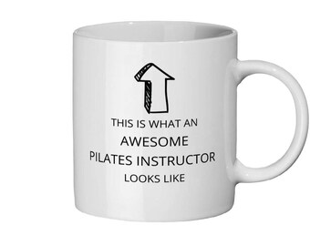 Op de afbeelding: Witte keramische mok met een zwarte pijlgrafiek en de tekst "THIS IS WHAT AN AWESOME PILATES INSTRUCTOR LOOKS LIKE". De mok heeft een gebogen handvat en is geschikt voor warme of koude dranken.