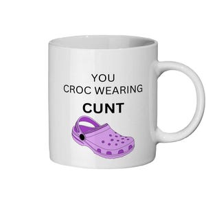 Può includere: Tazza in ceramica bianca con manico. La tazza presenta il testo "YOU CROC WEARING CUNT" sopra un'immagine a cartoni animati viola di una scarpa Croc.