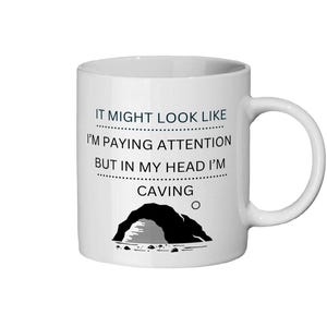 May include: White ceramic mug with a black handle. The mug features the text "IT MIGHT LOOK LIKE I'M PAYING ATTENTION BUT IN MY HEAD I'M CAVING" in black, along with a black cave illustration.