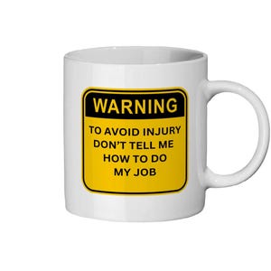 May include: White ceramic mug with a black and yellow warning label. The label reads "WARNING TO AVOID INJURY DON'T TELL ME HOW TO DO MY JOB". The mug has a curved handle.