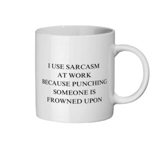 May include: White ceramic mug with a black text that reads: "I USE SARCASM AT WORK BECAUSE PUNCHING SOMEONE IS FROWNED UPON." The mug has a standard handle and a cylindrical shape.