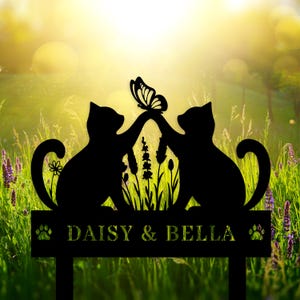 Op de afbeelding: Zwarte metalen tuinpaal met twee kat silhouetten en een vlinder. De paal heeft de namen "DAISY & BELLA" en pootafdrukken. Het ontwerp staat tegen een achtergrond van groen gras en paarse bloemen.