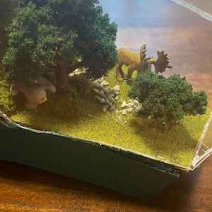 Puede incluir: Un diorama en miniatura que presenta una escena forestal con una figura de alce marrón, árboles verdes y un pájaro pequeño. La escena está ambientada sobre un lecho de musgo verde y pequeñas rocas, encerrada en un recipiente transparente.