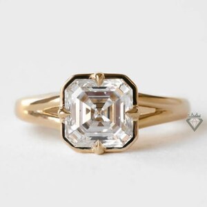 Peut inclure: Une bague en or ornée d'un grand diamant de taille carrée avec une bordure noire. La bague a un anneau simple. Les facettes du diamant reflètent la lumière. La bague est posée sur un fond blanc.