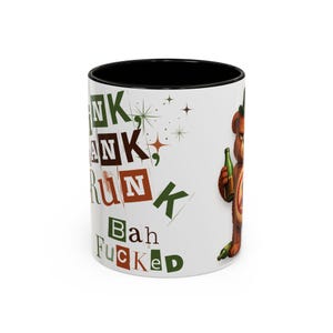 Op de afbeelding: Witte keramische mok met een zwarte binnenkant. De mok toont de woorden "Drunk, Run, Bah, Fucked" in verschillende lettertypen en kleuren. Een cartoonbeer met een groene fles staat rechts. De mok is een standaard formaat van ongeveer 325 ml.