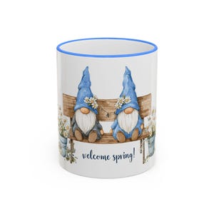 May include: White ceramic mug with a blue rim featuring two watercolor gnomes in blue hats sitting on a wooden bench. The gnomes are decorated with daisies. The words "welcome spring!" are written in a script font.