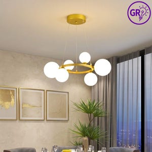 Puede incluir: Una lámpara de araña dorada moderna con seis pantallas esféricas de vidrio blanco. La lámpara tiene un diseño circular con un acabado dorado y está suspendida del techo. La habitación presenta obras de arte enmarcadas y una planta.