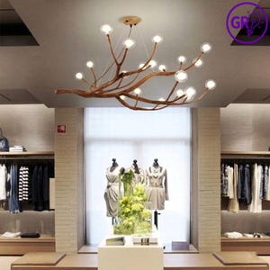 Puede incluir: Una lámpara de araña moderna con un diseño de rama de madera natural. La lámpara cuenta con múltiples bombillas pequeñas, redondas y transparentes. La lámpara de araña cuelga en una tienda minorista con percheros y maniquíes.